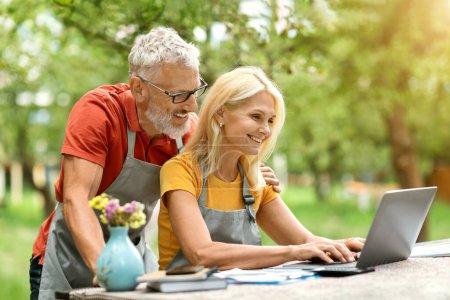 Photo for Happy Mature Spouses Using Laptop Computer While Sitting At Garden Terrace, Smiling Senior Couple Wearing Aprons Browsing Internet, Making Online Shopping Together, Woman Typing On Keyboard - Royalty Free Image