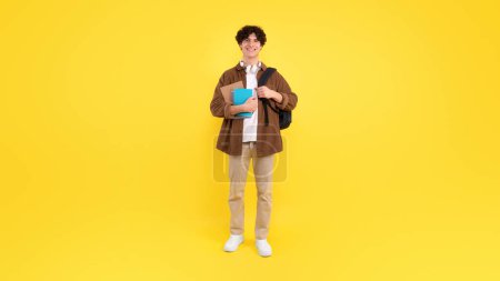 Photo for University Education concept. Positive young man with backpack, notebooks and headphones posing standing on yellow studio background, smiling to camera. Full length shot of student, panorama - Royalty Free Image