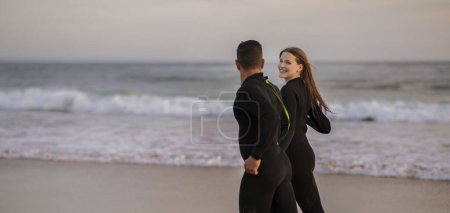 Téléchargez les photos : Couple de surfeurs souriants heureux courant ensemble le long de la plage, Jeune homme et femme romantique portant des combinaisons de surf ensemble sur la côte de l'océan, Profitant des sports nautiques, Panorama avec espace de copie - en image libre de droit