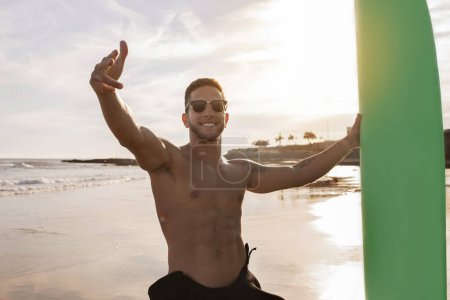 Photo for Portrait Of Young Handsome Male Surfer Posing With Surfboard On The Beach At Sunset Time, Smiling Sporty Millennial Guy In Stylish Sunglasses Pointing Fingers At Camera, Enjoying Surfing, Free Space - Royalty Free Image