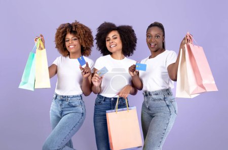 Foto de Tres clientes sonrientes Damas negras con bolsas de compras que muestran tarjetas de crédito, oferta de venta de publicidad y pagos bancarios fáciles, de pie en el fondo del estudio púrpura. Concepto de compras exitosas - Imagen libre de derechos