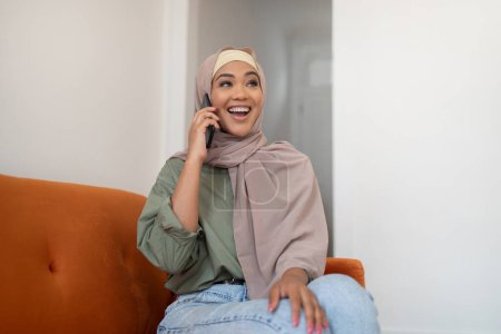 Téléchargez les photos : Communication mobile. Joyeuse dame noire dans le hijab parlant sur un téléphone portable assis sur un canapé à la maison, appréciant la conversation, communiquant avec des amis via un téléphone intelligent, regardant à côté - en image libre de droit
