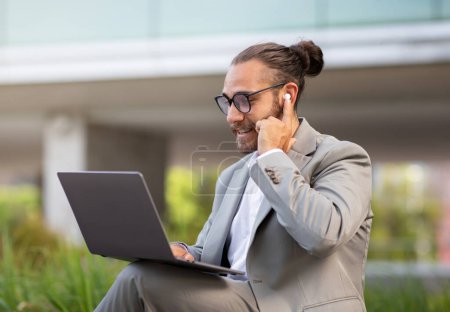 Foto de Hombre de negocios milenario guapo haciendo videollamada en el ordenador portátil mientras está sentado al aire libre, joven gerente sonriente en traje trabajando fuera de la oficina, disfrutando de la comunicación en línea y las tecnologías modernas - Imagen libre de derechos