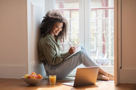 Photo for Online Study And Work. Happy Black Woman Using Laptop Taking Notes In Notebook, Sitting On Floor Near Window At Home, Learning Online And Writing Article. Freelancer Lifestyle, Internet Education - Royalty Free Image