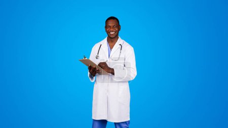Photo for Smiling handsome young african american doctor in white coat making notes in clipboard, enjoy work, isolated on blue studio background. Exam of patient, medicine, treatment in clinic, health care - Royalty Free Image