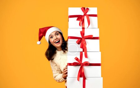Téléchargez les photos : Cadeau de Noël. Joyeux jeune femme dans le chapeau rouge Santa tenant de nombreux cadeaux boîtes souriant à la caméra posant sur fond jaune, Studio Shot. Vacances d'hiver, Noël présente l'offre de livraison - en image libre de droit