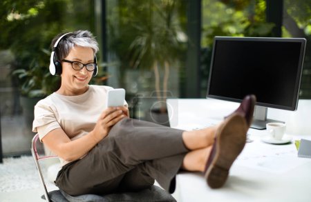 Photo for Relaxed happy mature woman in smart casual employee have break at home office, sitting at workplace with legs on desk, using smartphone, wireless headphones, listening to music, watching video podcast - Royalty Free Image