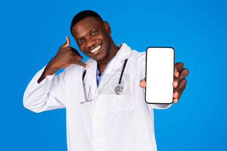 Téléchargez les photos : Heureux jeune médecin afro-américain en manteau montre smartphone avec écran blanc, appelez-moi signe avec la main, isolé sur fond de studio bleu. Communication, application, recommandation de site Web pour les soins de santé - en image libre de droit