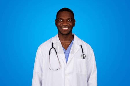 Photo for Positive young black guy doctor in white coat enjoy work, isolated on blue studio background. Professional occupation, health care, medical services in clinic, exam and treatment - Royalty Free Image