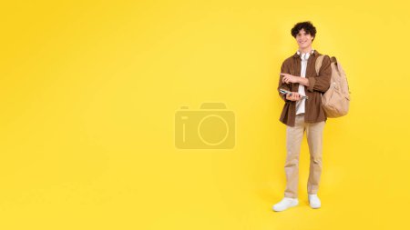 Photo for Happy caucasian young man smiling and pointing aside at free space, holding his notebooks and backpack on yellow studio background, full length shot of student advertising education offer, panorama - Royalty Free Image
