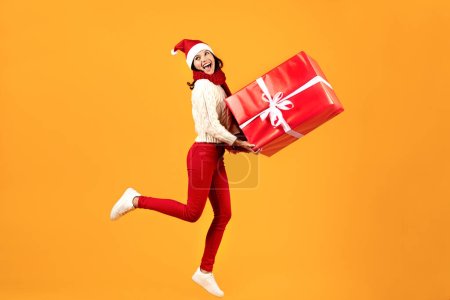 Photo for Christmas Gift Delivery. Joyful Young Woman Posing With Big Wrapped Present Box, Wearing Red Santa Hat, Advertising Xmas Winter Offer, Jumping In Mid Air On Yellow Background In Studio - Royalty Free Image