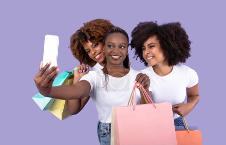 Téléchargez les photos : Black Friday Selfie. Trois femmes afro-américaines dentelées avec des sacs à provisions posant prenant des photos sur le téléphone, s'amusant tout en achetant des vêtements sur fond violet. Saison de vente dans le centre commercial - en image libre de droit