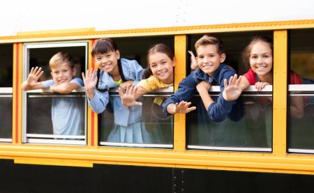 Foto de Grupo de niños felices asomándose por la ventana del autobús escolar, saludando y sonriendo a la cámara, niños y niñas alegres disfrutando de su viaje en autobús escolar amarillo, transporte para el concepto de los alumnos - Imagen libre de derechos