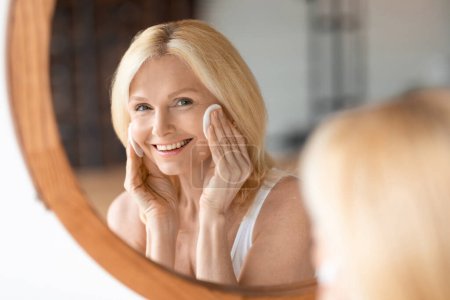 Foto de Hermosa mujer madura caucásica mirando el espejo y limpiando la piel con almohadillas de algodón, haciendo rutina de belleza diaria en casa. Mujer disfrutando de tratamientos para el cuidado de la piel - Imagen libre de derechos
