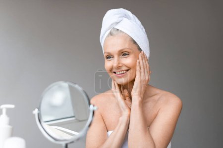Téléchargez les photos : Belle femme mature avec serviette sur la tête regardant le miroir, touchant sa peau du visage et souriant, en utilisant de nouveaux cosmétiques anti-âge de marque, intérieur de la maison, espace libre - en image libre de droit