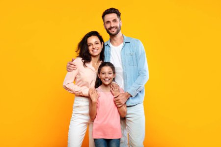 Téléchargez les photos : Heureuse famille européenne de trois câlins et souriant à la caméra, posant ensemble sur fond de studio jaune. Mère et père embrassant leur jolie fille - en image libre de droit