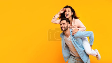 Photo for Young couple european man giving piggyback ride to joyful wife sitting on back and looking away, holding hand at forehead, yellow background, banner - Royalty Free Image