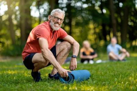 Téléchargez les photos : Beau senior homme roulant tapis de yoga après la leçon de groupe à l'extérieur, heureux homme mûr en vêtements de sport regardant la caméra, profiter des séances d'entraînement à l'extérieur et mode de vie actif à la retraite, foyer sélectif - en image libre de droit