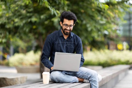 Photo for Cheerful handsome young hindu guy businessman chatting with client, using laptop at park, drink takeaway coffee, work outdoors. Independent contractor checking emails, look for new job opportunities - Royalty Free Image