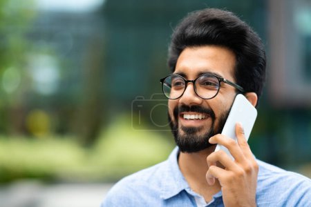 Foto de Retrato de primer plano del alegre guapo empresario indio milenario que usa anteojos hablando por teléfono con su pareja de negocios o cliente mientras camina al aire libre, mirando el espacio de copia y sonriendo - Imagen libre de derechos