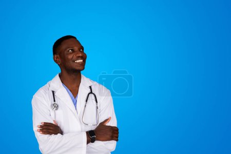 Photo for Positive young african american doctor in white coat with crossed arms on chest looks at copy space, isolated on blue studio background. Dream, health care, medicine in clinic, ad and offer - Royalty Free Image