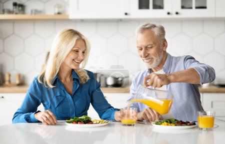 Téléchargez les photos : Heureux aînés conjoints manger savoureux sain petit déjeuner et boire du jus d'orange dans la cuisine, joyeux couple d'âge mûr profiter de délicieux aliments ensemble à la maison, vieil homme versant boisson à la femme - en image libre de droit