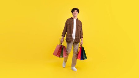 Photo for Big sale and shopping leisure. Cheerful young guy holding many paper bags smiling to camera on yellow background in studio. Panorama, full length of contented buyer with packages and purchases - Royalty Free Image