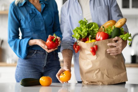 Photo for Cropped Shot Of Man And Woman Unpacking Paper Bag With Groceries In Kitchen At Home, Unrecognizable Mature Couple After Food Shopping Puting Fresh Organic Vegetables On Table, Closeup - Royalty Free Image