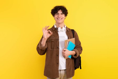 Téléchargez les photos : Succès du jeune étudiant homme avec des cahiers et des gestes de sac à dos ok, approuvant le succès éducatif, souriant à la caméra posant sur fond jaune en studio. Tout est OK concept - en image libre de droit