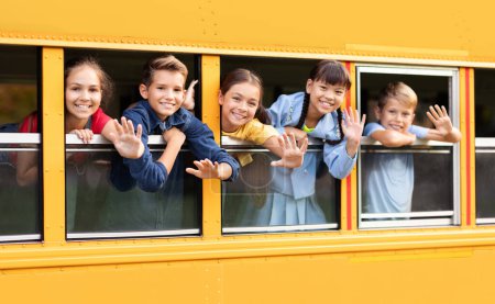Foto de Niños felices saludando con las manos mientras asoman por la ventana amarilla del autobús escolar, retrato de niños y niñas alegres que viajan juntos a la escuela, alumnos alegres sonriendo ante la cámara, primer plano - Imagen libre de derechos