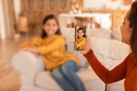 Foto de Compartiendo. Árabe joven madre haciendo foto de su linda hija preadolescente en el teléfono inteligente interior, salvando recuerdos y momentos felices de la familia, compartiendo la vida en línea en las redes sociales. Enfoque selectivo - Imagen libre de derechos