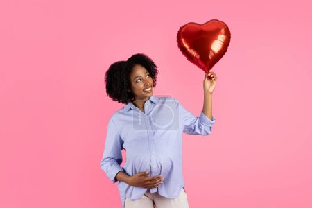 Foto de La señora embarazada negra milenaria positiva que toca el vientre grande, sostiene el globo inflable en forma de corazón, goza de maternidad futura, aislado en fondo rosado, estudio. Esperando fiesta infantil y amor - Imagen libre de derechos