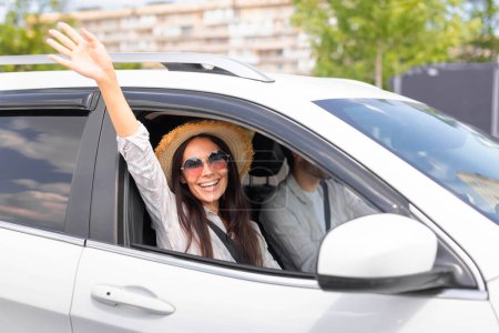 Téléchargez les photos : Jolie dame millénaire insouciante portant un chapeau en osier et des lunettes de soleil agitant la main et souriant à la caméra depuis la fenêtre ouverte de la voiture, une voyageuse profitant d'un voyage en voiture avec son petit ami ou son mari - en image libre de droit