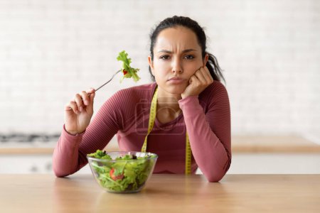 Foto de Mujer fitness infeliz adelgazando comiendo ensalada de verduras, sosteniendo tenedor y mirando a la cámara con expresión triste que sufre de falta de apetito, sentado en la mesa en la cocina moderna. Dietas luchas - Imagen libre de derechos