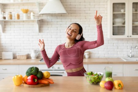 Photo for Healthy Weight Loss Lifestyle. Joyful Fit Woman Wearing Wireless Headphones, Dancing And Listening To Music Singing At Kitchen, Enjoying Slimming Routine Near Table With Healthy Vegetables - Royalty Free Image