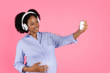 Photo for Glad millennial black lady with belly in wireless headphones listens to music, enjoys free time, makes selfie on phone, isolated on pink background, studio. Lifestyle, blog and social networks - Royalty Free Image