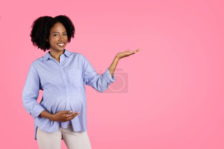 Photo for Positive millennial black pregnant lady touching big belly, hold copy space, enjoy motherhood, isolated on pink background, studio. Recommends for expecting child, sale, ad, offer - Royalty Free Image