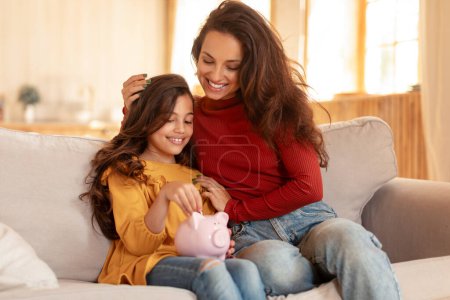Téléchargez les photos : Leçons de finances familiales. Arabe maman et fille partageant la joie d'économiser, mettre de l'argent dans une tirelire, investir dans un avenir financier pour les enfants, assis sur le canapé dans le salon moderne à l'intérieur - en image libre de droit