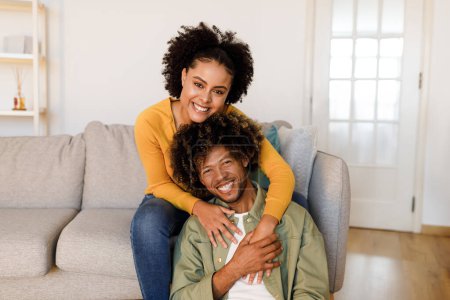 Téléchargez les photos : Heureux couple afro-américain partageant un étreinte chaude sur le canapé intérieur, épouse étreignant mari de retour dans leur salon confortable, souriant à la caméra, rayonnant d'amour et de bonheur ensemble - en image libre de droit