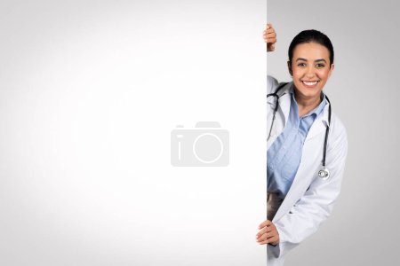 Téléchargez les photos : Publicité médicale. Joyeuse médecin latino-américaine regardant hors du grand panneau publicitaire vierge, joyeuse thérapeute brésilienne en uniforme démontrant l'espace vide sur une pancarte blanche, panorama - en image libre de droit
