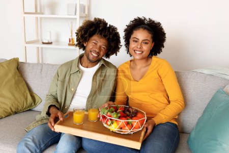 Téléchargez les photos : Nutrition familiale saine. Heureux couple afro-américain assis tenant plateau avec des fruits frais et du jus, profiter du temps de collation sur le canapé à la maison, sourire à la caméra. Vitamines, régime alimentaire stimulant l'immunité - en image libre de droit