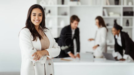Foto de Empresario moderno. Joven mujer de negocios brasileña posando en la sala de conferencias en la oficina durante la reunión con colegas, mujer de negocios latina de pie con los brazos cruzados y sonriendo - Imagen libre de derechos