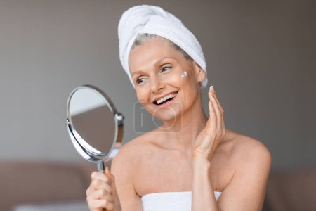 Téléchargez les photos : Belle femme mûre souriante utilisant une crème hydratante pour le visage, regardant le miroir, appréciant sa routine de soins du visage du matin, assise dans l'intérieur de la chambre - en image libre de droit