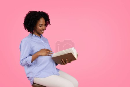 Photo for Positive millennial black pregnant lady touch belly with hand, read book, rest, enjoy motherhood, isolated on pink background, studio. Expecting child, health care, hobbies, study in free time - Royalty Free Image