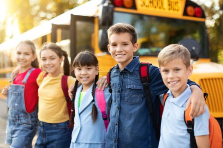 Foto de Niños multiétnicos felices posando al aire libre cerca del autobús escolar, niños y niñas preadolescentes alegres con mochilas afuera, abrazando y sonriendo en la cámara, disfrutando de estudio juntos, primer plano - Imagen libre de derechos
