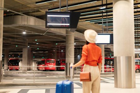 Photo for Tourist Woman Standing With Travel Suitcase At Bus Station And Looking At Departure Board, Back View Shot Of Unrecognizable Passenger with Luggage At Terminal. Selective Focus - Royalty Free Image