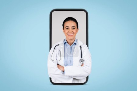 Foto de Joven doctora brasileña en uniforme blanco posando con estetoscopio y brazos cruzados en enorme pantalla de teléfono celular, fondo azul, pancarta. Concepto de servicios médicos - Imagen libre de derechos