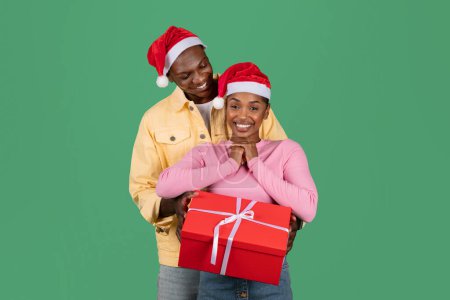 Photo for Happy holidays. Handsome black guy hugging from behind and giving red gift box to his excited beautiful young african american girlfriend wife, loving couple celebrating xmas on green background - Royalty Free Image