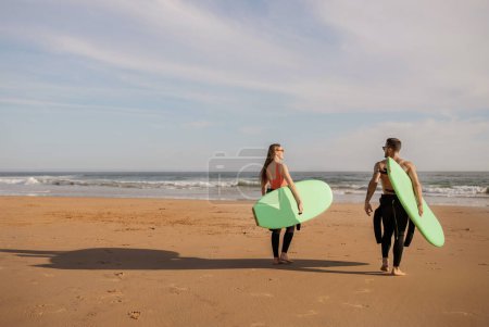 Foto de Pareja joven de surfistas caminando en la orilla del mar con tablas de surf, feliz joven millennial hombre y mujer en trajes de neopreno que van al océano, listos para surfear juntos, disfrutando de deportes acuáticos, espacio de copia - Imagen libre de derechos