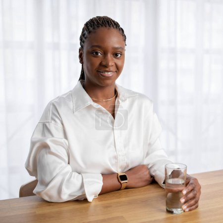Foto de Retrato de una atractiva joven empresaria negra en traje formal sentada en el escritorio, bebiendo agua dulce en la oficina, mirando a la cámara y sonriendo, espacio para copiar. Hidratación, concepto de estilo de vida saludable - Imagen libre de derechos